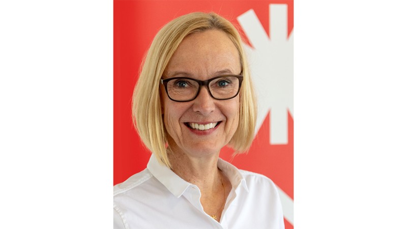 Portrait of a smiling woman with blonde hair and glasses in front of a red background with a white symbol.