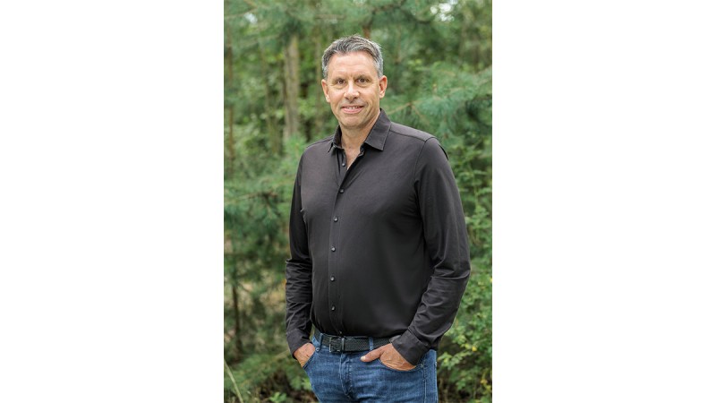 Man smiling outdoors in front of green forest, wearing a dark shirt and jeans.