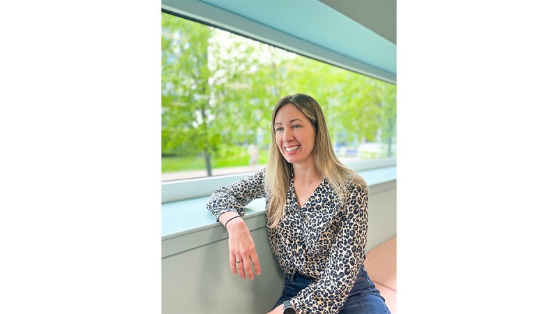 Smiling woman in a leopard print blouse sitting by a window overlooking greenery.