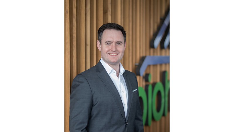 Man in a suit smiling at the camera, standing in front of a wooden wall with a green logo.