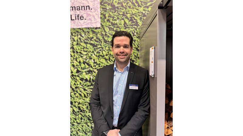 Man in a suit smiling in front of a trade fair wall with a leaf pattern, wearing a name badge.