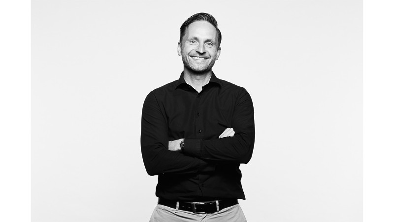 Man with crossed arms smiling in a black and white portrait against a light background.