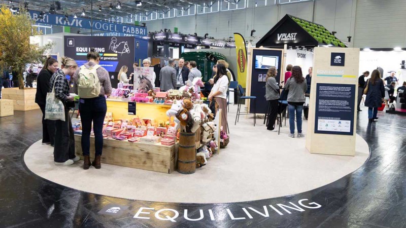 Trade show booth with 'EQUI LIVING' written on the floor, several people looking at products displayed on wooden tables and barrels.